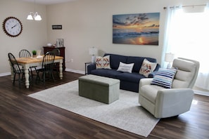 Un salon avec un canapé bleu foncé, un fauteuil blanc, une table à manger en bois avec des chaises, un panneau indiquant un bar à café et un tableau représentant un coucher de soleil accroché au mur.