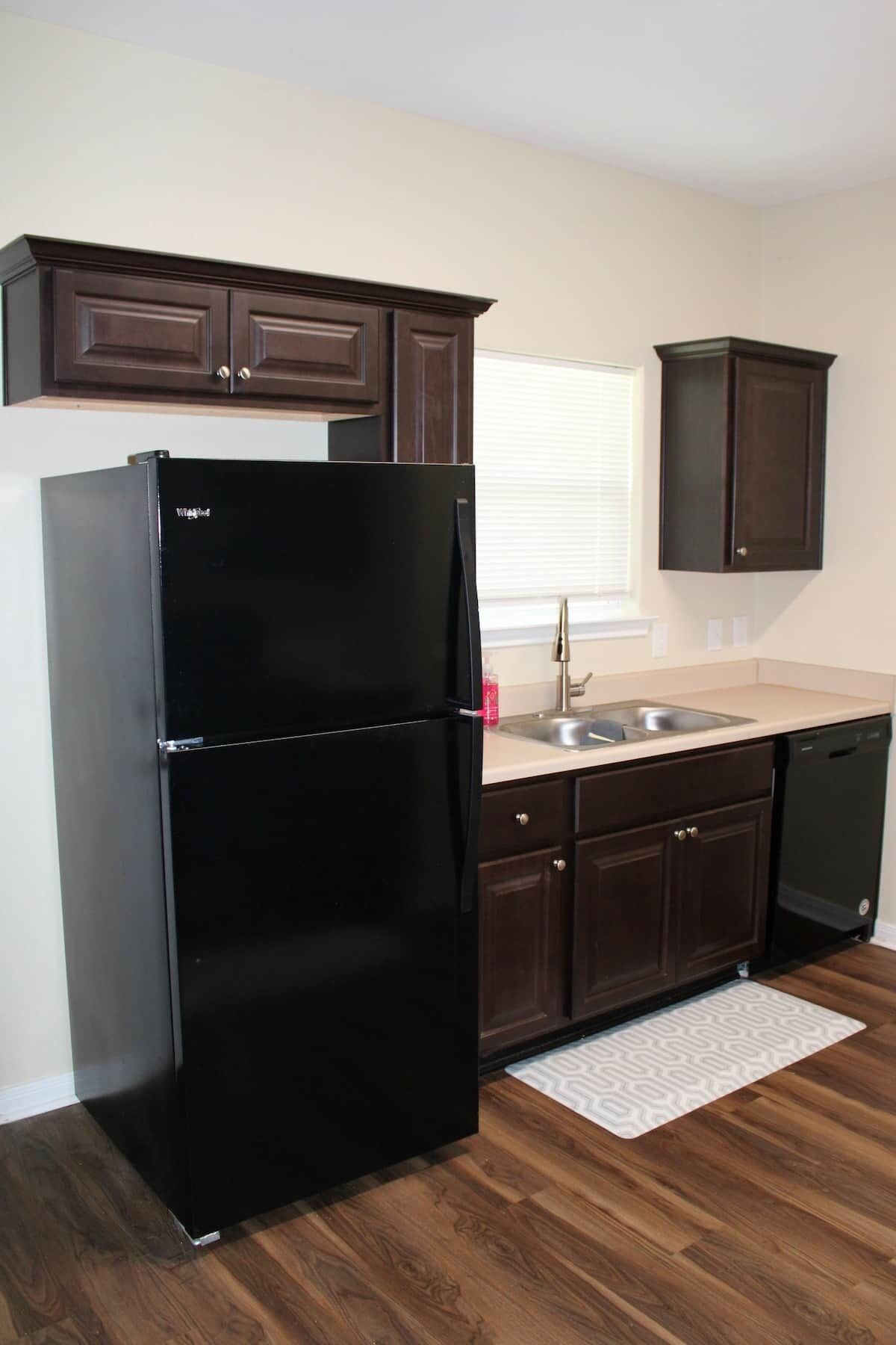 A kitchen with dark wooden cabinets, a black refrigerator, a dishwasher, and a white countertop.