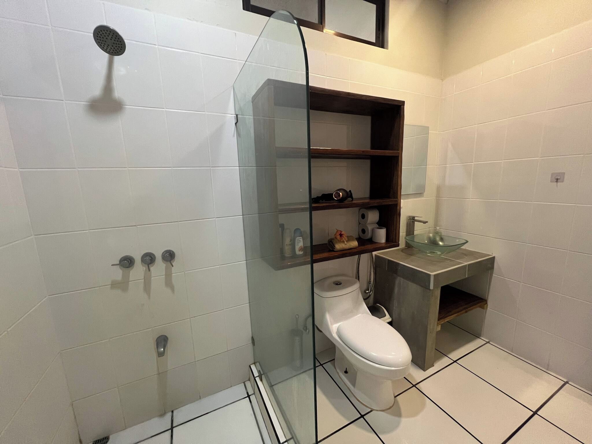 A modern bathroom with a glass-enclosed shower, a toilet, a sink with a glass bowl, and a shelf with toiletries.