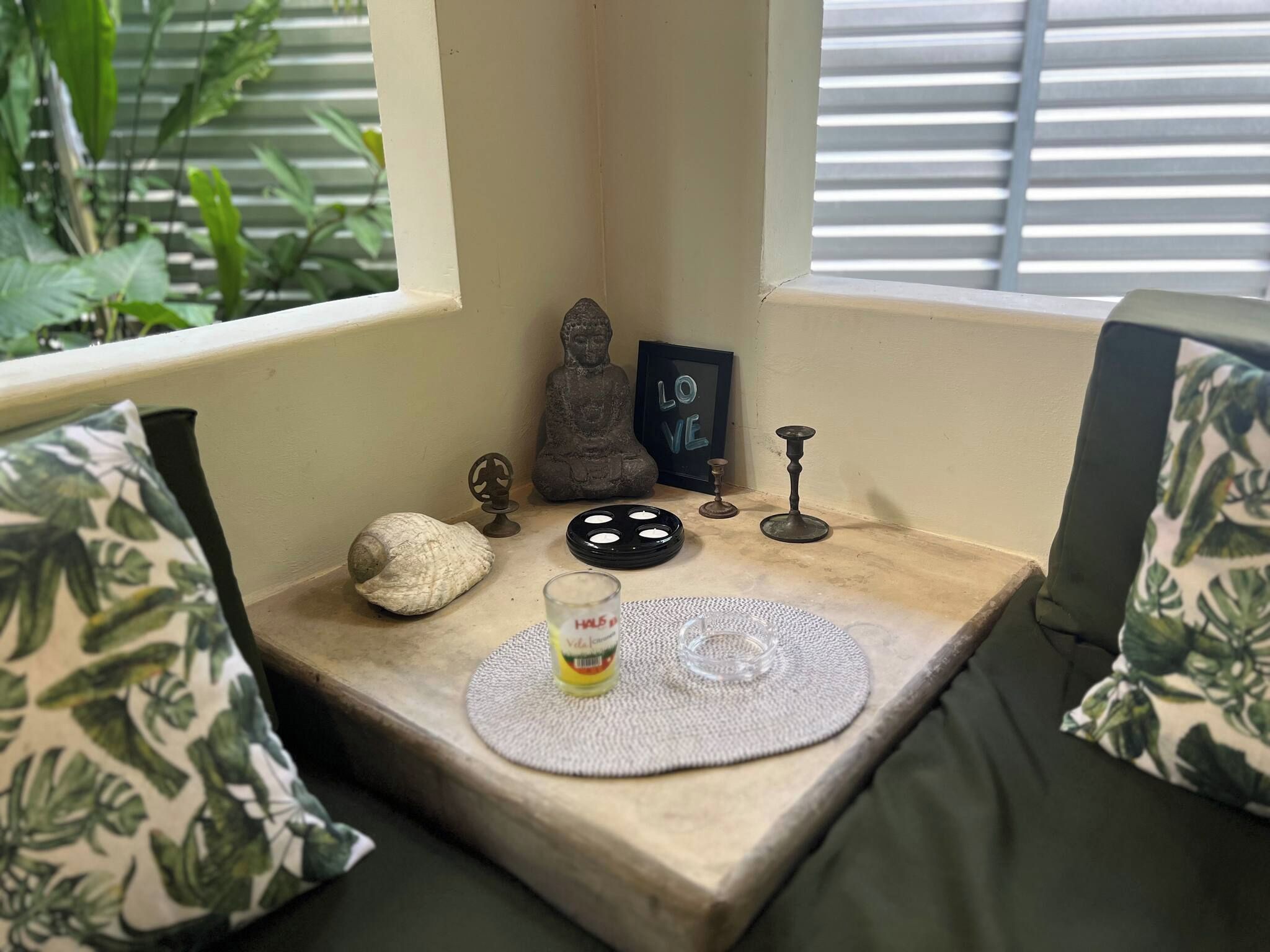 A cozy corner with a cushioned bench, a small table with a mat, a Buddha statue, a framed sign, a candle holder, and a decorative shell.