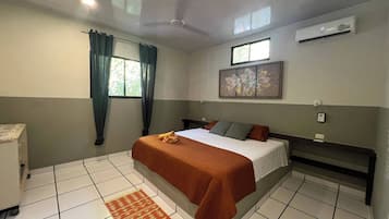 Habitación de hotel con cama, ventana con cortinas, ventilador de techo, aire acondicionado y suelo de baldosas.