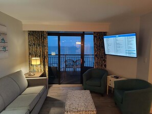 Une pièce comprenant un canapé, deux fauteuils, une petite table, un téléviseur affichant une liste de chaînes, et un balcon avec vue sur la mer.