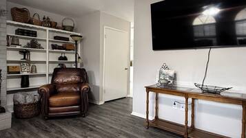 A living room with a leather armchair, a wooden coffee table, a bookshelf, and a mounted flat-screen TV.