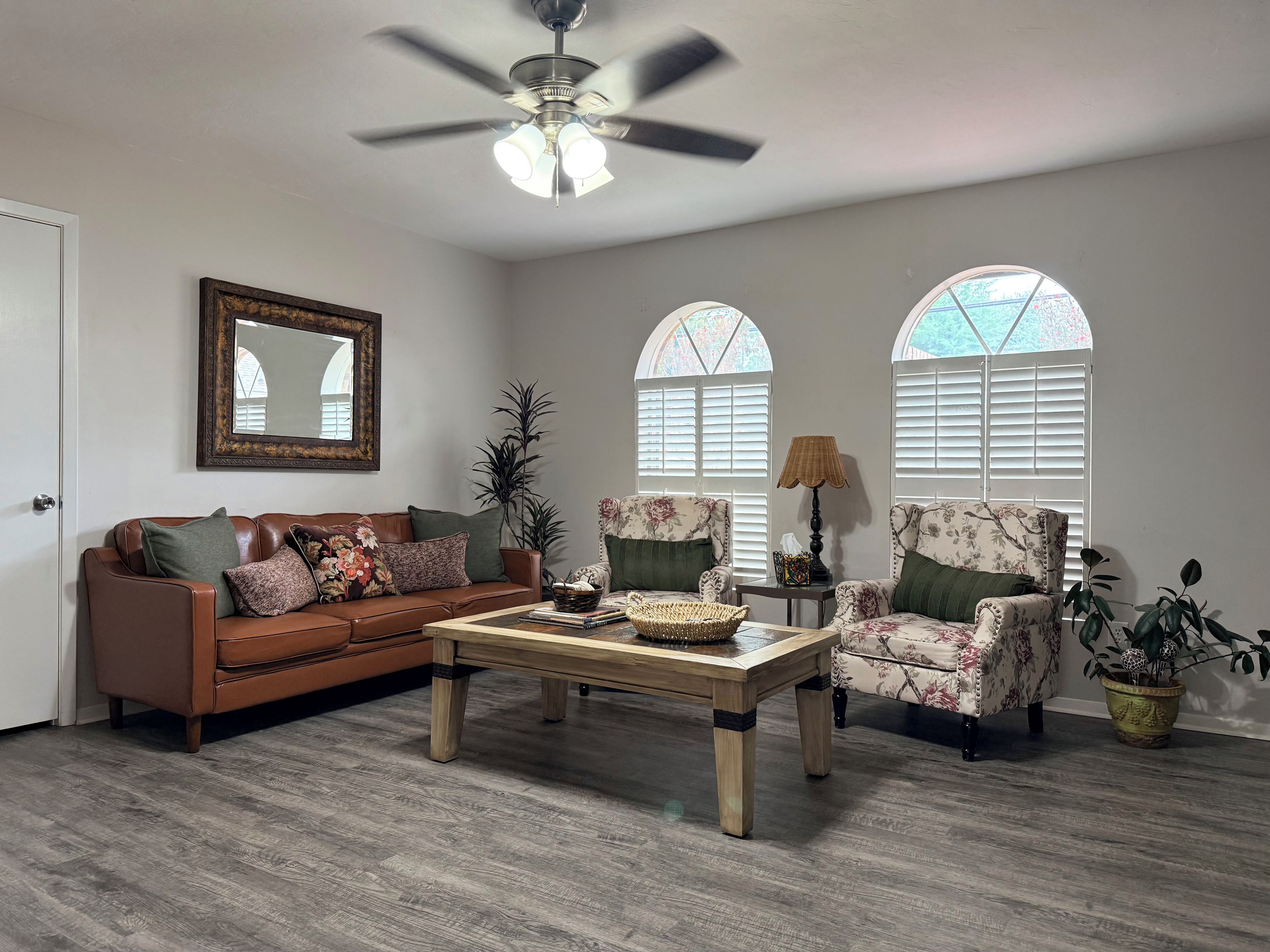 A living room with a brown leather sofa, a floral armchair, a wooden coffee table, and a ceiling fan.