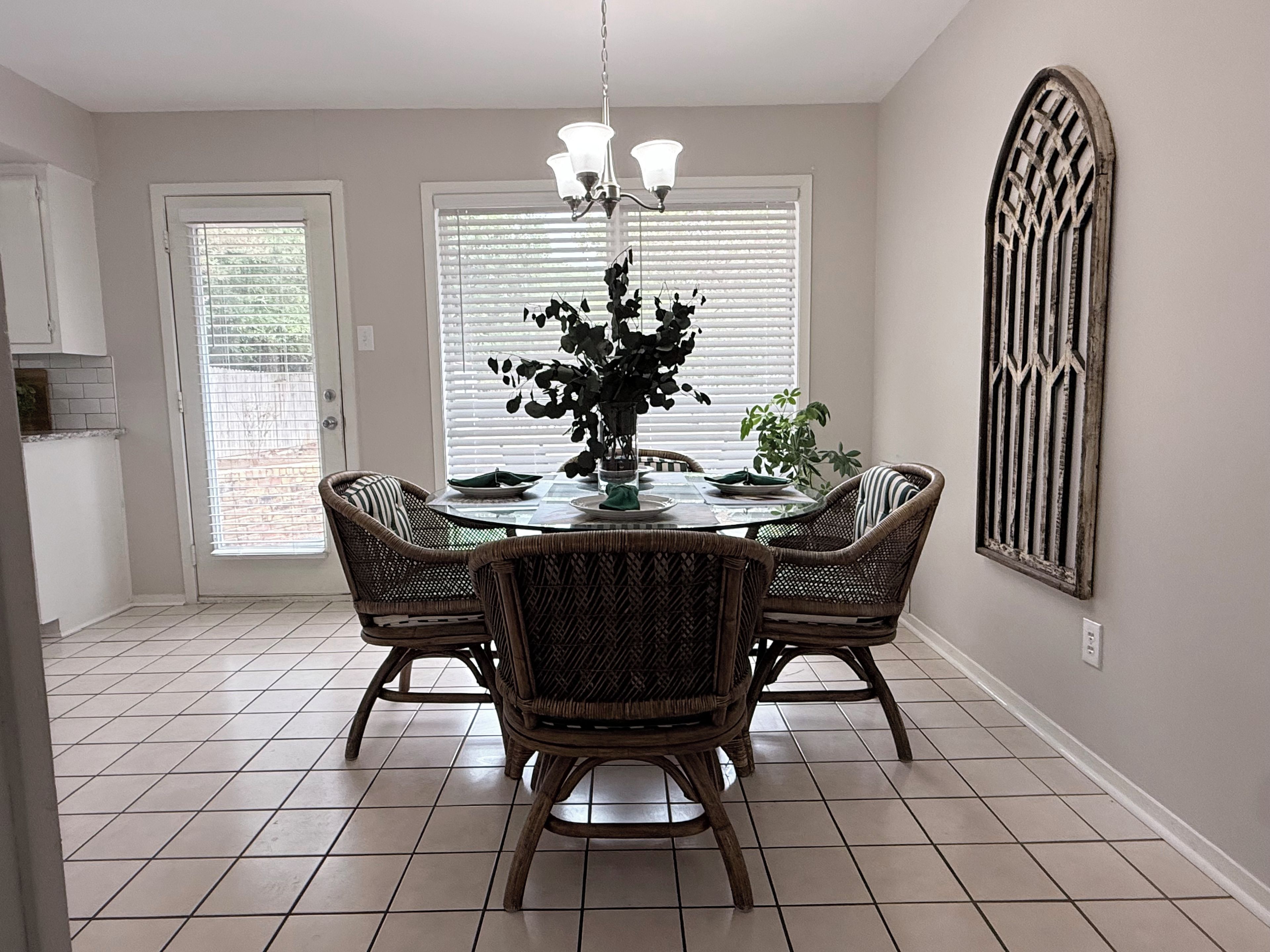 A dining area with a round table, four wicker chairs, a chandelier, and a decorative wall piece.
