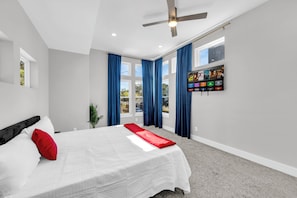 A modern bedroom with a bed, a ceiling fan, a TV mounted on the wall, and large windows with curtains. - Cozy 3BR - Walk to Fair Park - Pool Table, Fire Pit (Dallas)