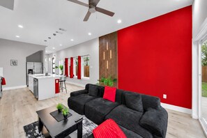 A modern kitchen with a central island, a dining area with red curtains, a black sofa with red cushions, and a ceiling fan. - Cozy 3BR - Walk to Fair Park - Pool Table, Fire Pit (Dallas)