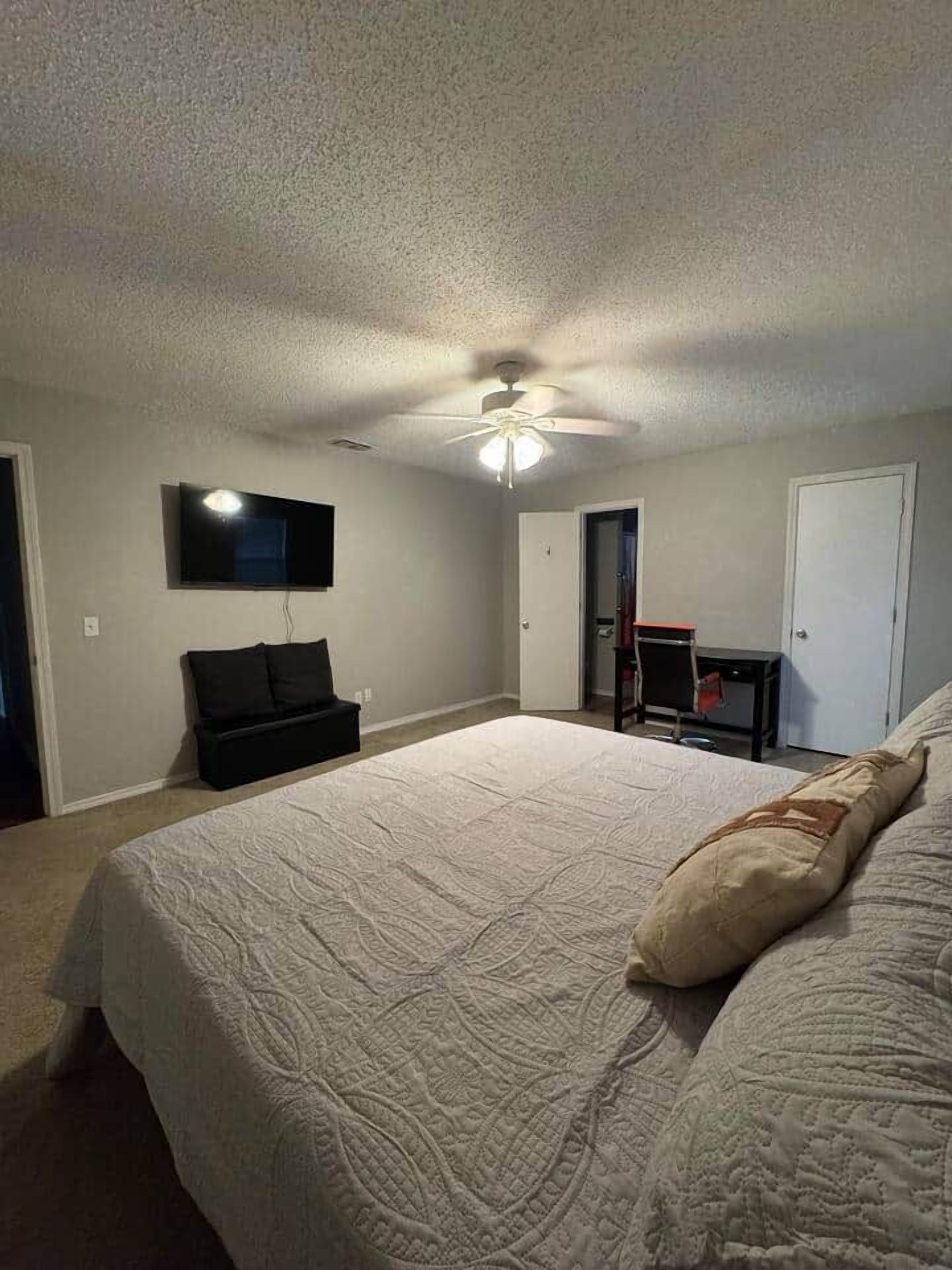 Quarto com cama, ventilador de teto, TV fixada na parede e porta que dá para outro cômodo.