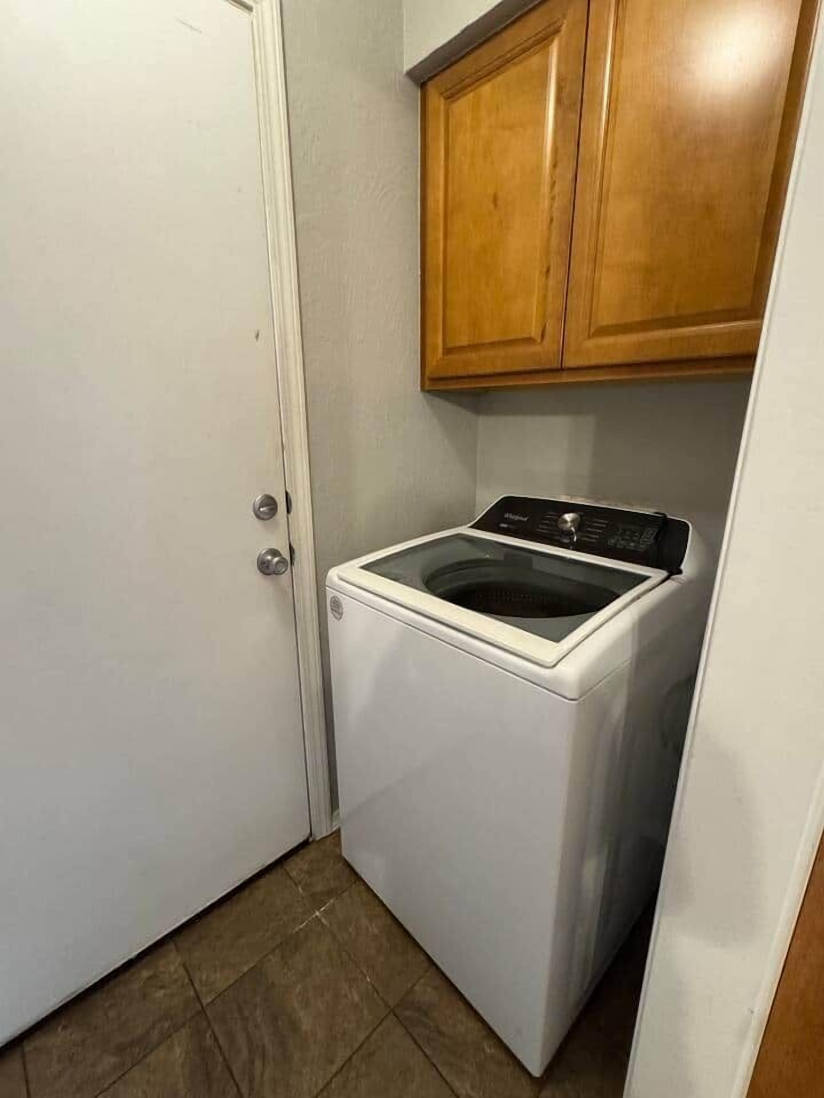 A compact laundry room with a front-loading washer and dryer, wooden cabinets above, and a white door.