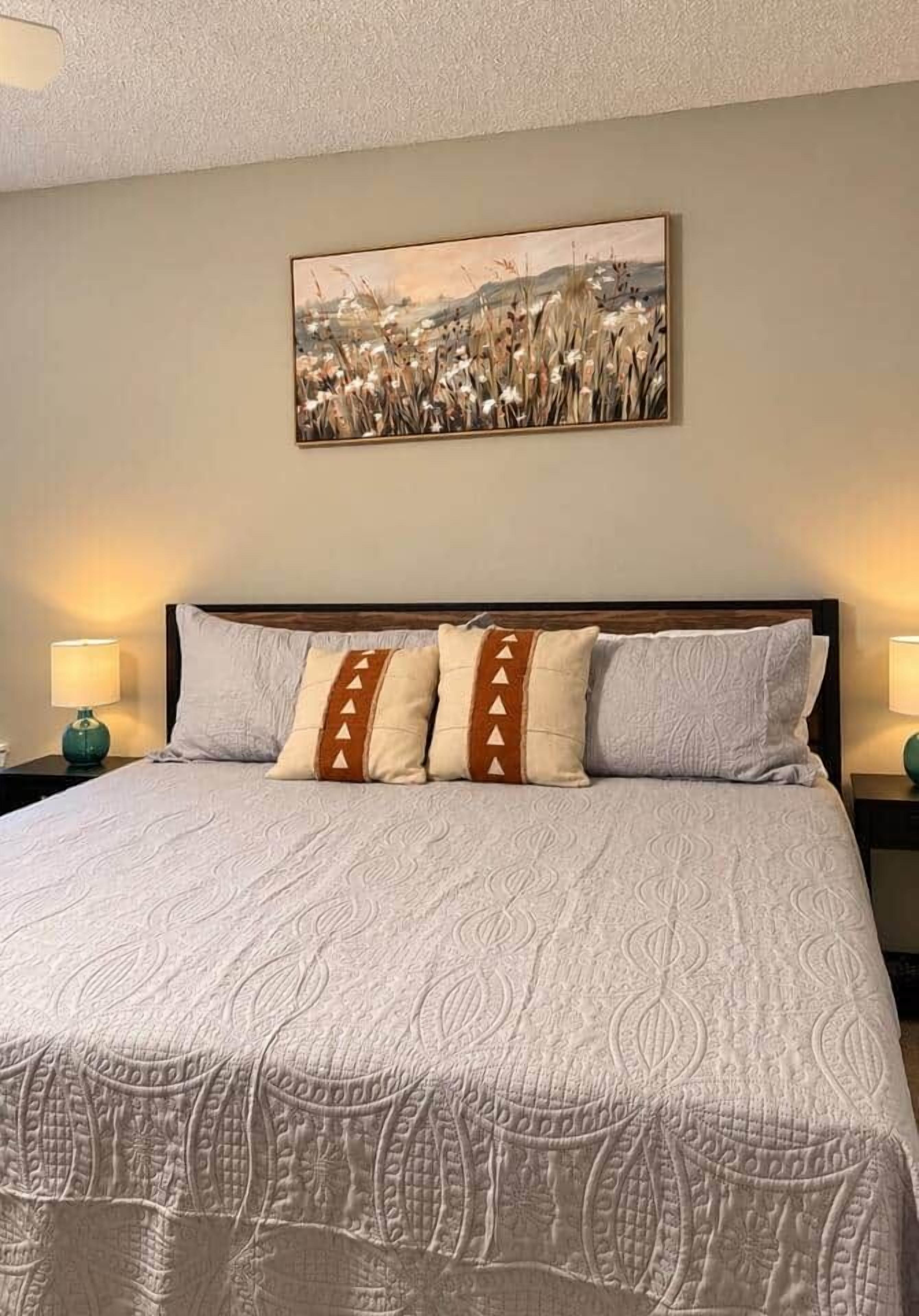 A neatly made bed with a patterned bedspread and decorative pillows, a painting above the bed, and two bedside lamps on either side.