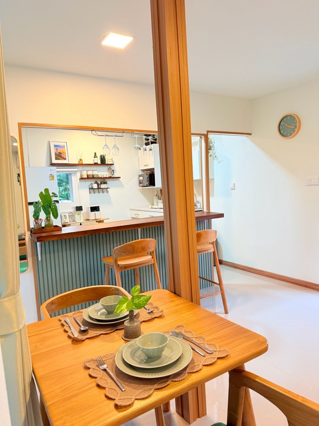 Un comedor con una mesa de madera preparada para dos, una cocina al fondo y un reloj en la pared.