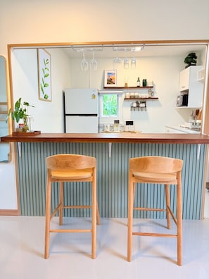 A kitchen with a wooden bar counter, two wooden bar stools, a refrigerator, a microwave, and a window with a view of greenery. - Curve House Lanta (Ko Lanta)