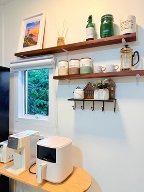 Un área de cocina con cafetera, una ventana que ofrece vistas a la vegetación y estantes con utensilios de cocina y objetos decorativos.