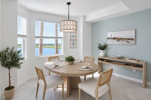 A dining area with a round table, chairs, a hanging light fixture, a view of the outdoors, and a wall-mounted shelf with decorative items. - Stunning Lakefront Calusa Country Club Condo - A Slice Of Paradise (Bradenton)