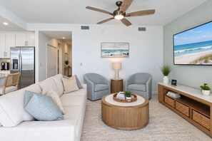 A modern living room with a beige sofa, two grey armchairs, a wooden coffee table, and a television displaying a beach scene. - Stunning Lakefront Calusa Country Club Condo - A Slice Of Paradise (Bradenton)