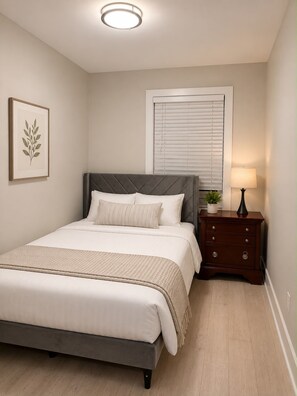 A bedroom with a bed, a bedside table with a lamp, a window with blinds, and a framed picture on the wall. - Modern 6BR Home Walk to Metro, Navy Yard & Wharf (Washington)