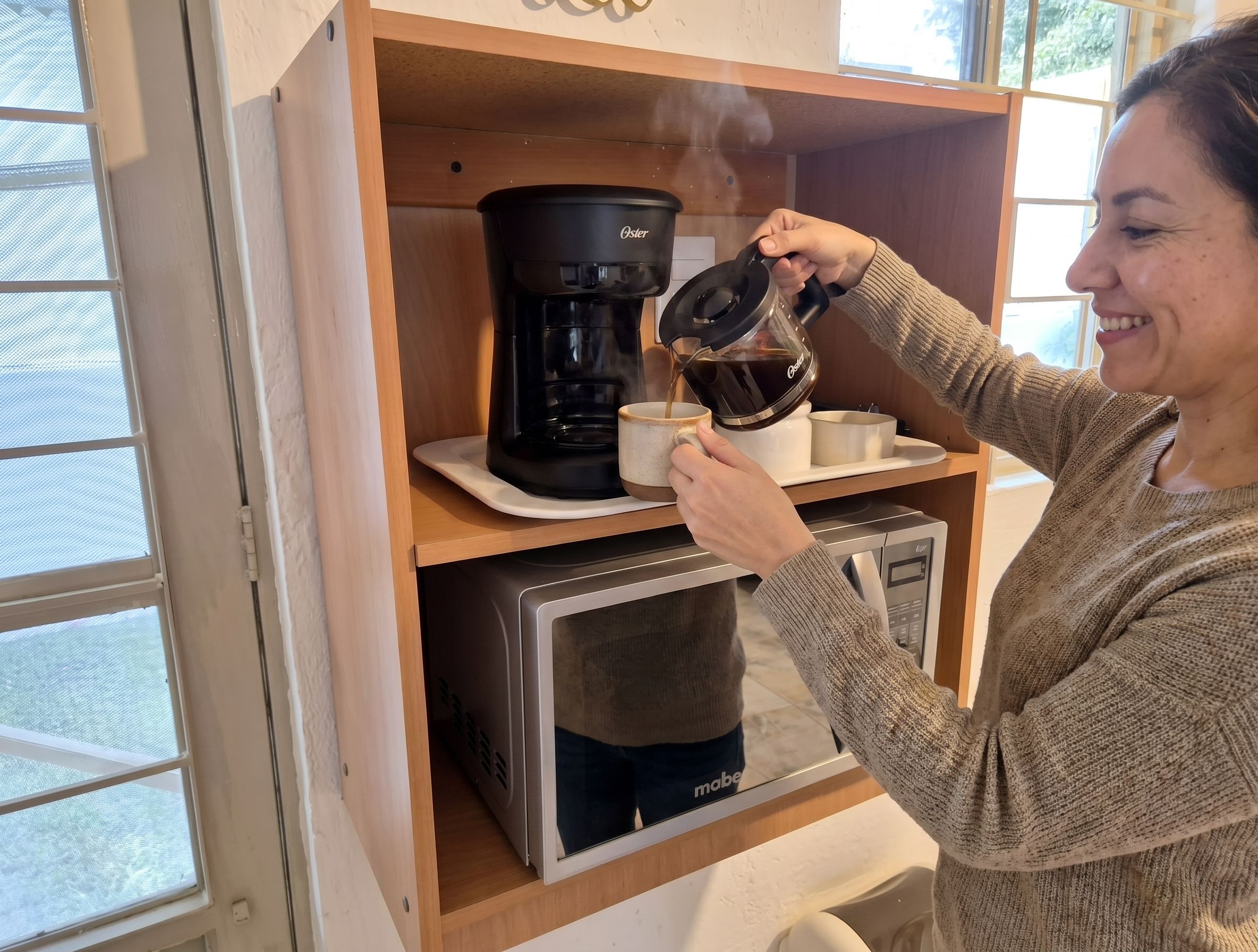 Iemand giet koffie in een mok vanuit een waterkoker, met een koffiezetapparaat en een magnetron eronder.