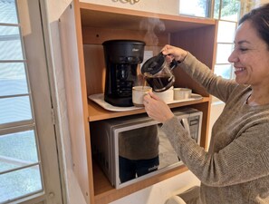 Iemand giet koffie in een mok vanuit een waterkoker, met een koffiezetapparaat en een magnetron eronder.