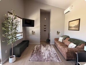 Ein Wohnzimmer mit einer Couch, einem Couchtisch, einer Pflanze und einem an der Wand montierten Fernseher.