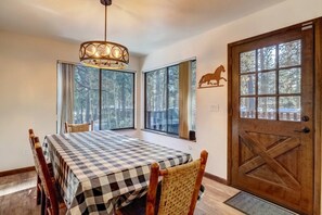 A dining area with a checkered table, wooden chairs, a chandelier, large windows with a view of trees, and a wooden door with a horse decoration. - Singing Pines - 4 bed, 4 bath Marla Bay home SLT (Glenbrook)