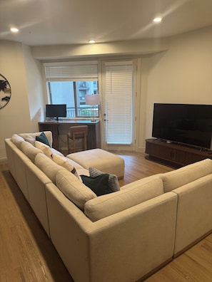 Ein Wohnzimmer mit einem Ecksofa, einem Fernseher, einem Schreibtisch mit Computer und einem Fenster mit Jalousien.