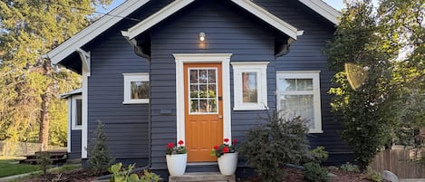 A two-story house with a front porch, a wooden door, and a small garden with potted plants.