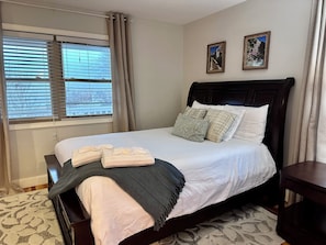 A neatly made bed with pillows and a blanket, a window with blinds, and two framed pictures on the wall.