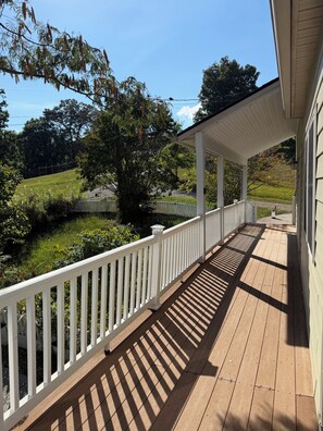 A covered porch with a white railing, wooden flooring, and a view of a green landscape.