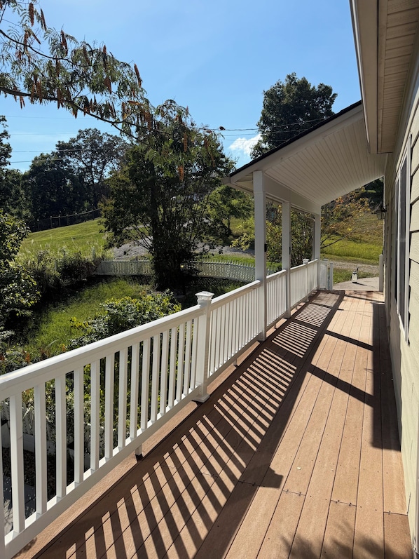 A covered porch with a white railing, wooden flooring, and a view of a green landscape.