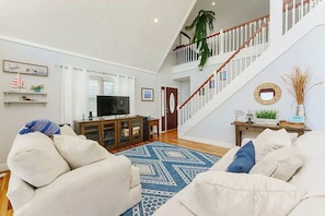 A spacious living room with a high ceiling, a staircase with white railings, a flat-screen TV on a wooden cabinet, and a patterned rug.