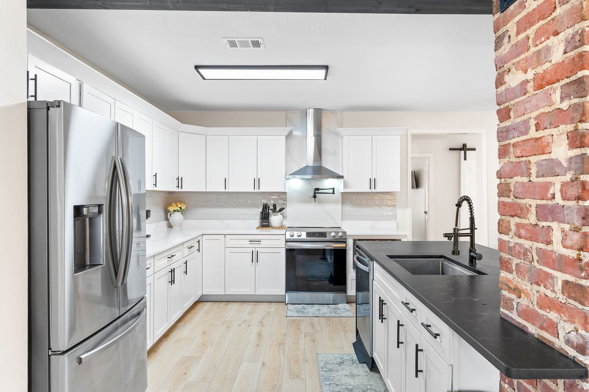 A modern kitchen with white cabinets, stainless steel appliances, a brick accent wall, and a black countertop.