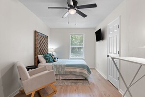 Ein Schlafzimmer mit einem Bett, einem Sessel, einem Deckenventilator, einem Fenster mit Jalousien und einem an der Wand befestigten Fernseher.