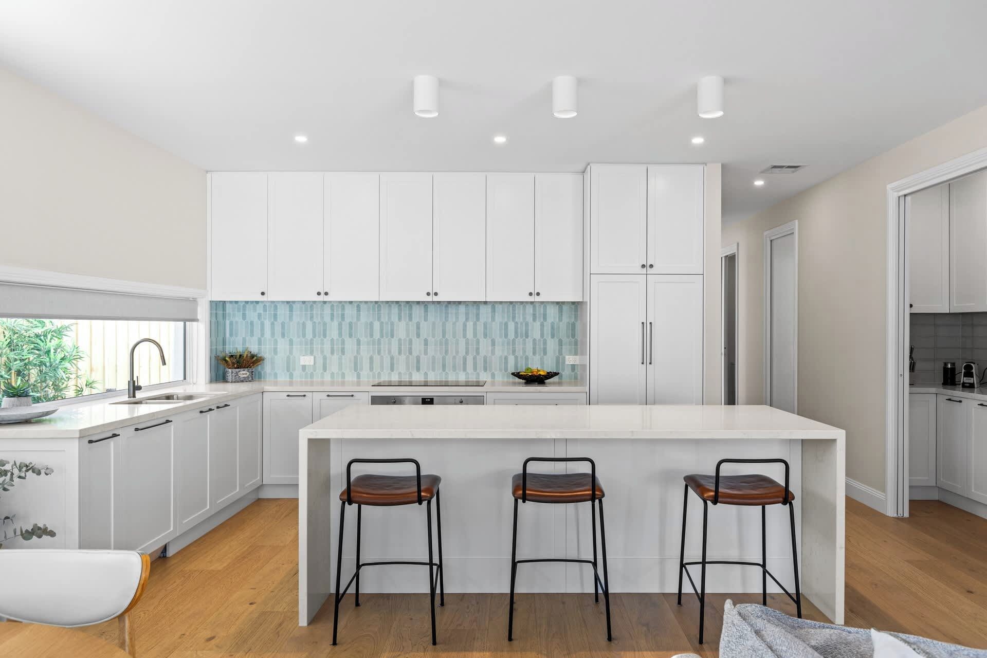 A modern kitchen with white cabinetry, a central island with bar stools, and a backsplash with blue tiles.