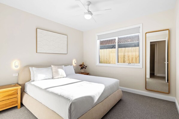A bedroom with a bed, a nightstand, a wall-mounted mirror, a ceiling fan, and a window with a view of a wooden fence. - Heated pool, Rosewood Retreat (Rosebud)