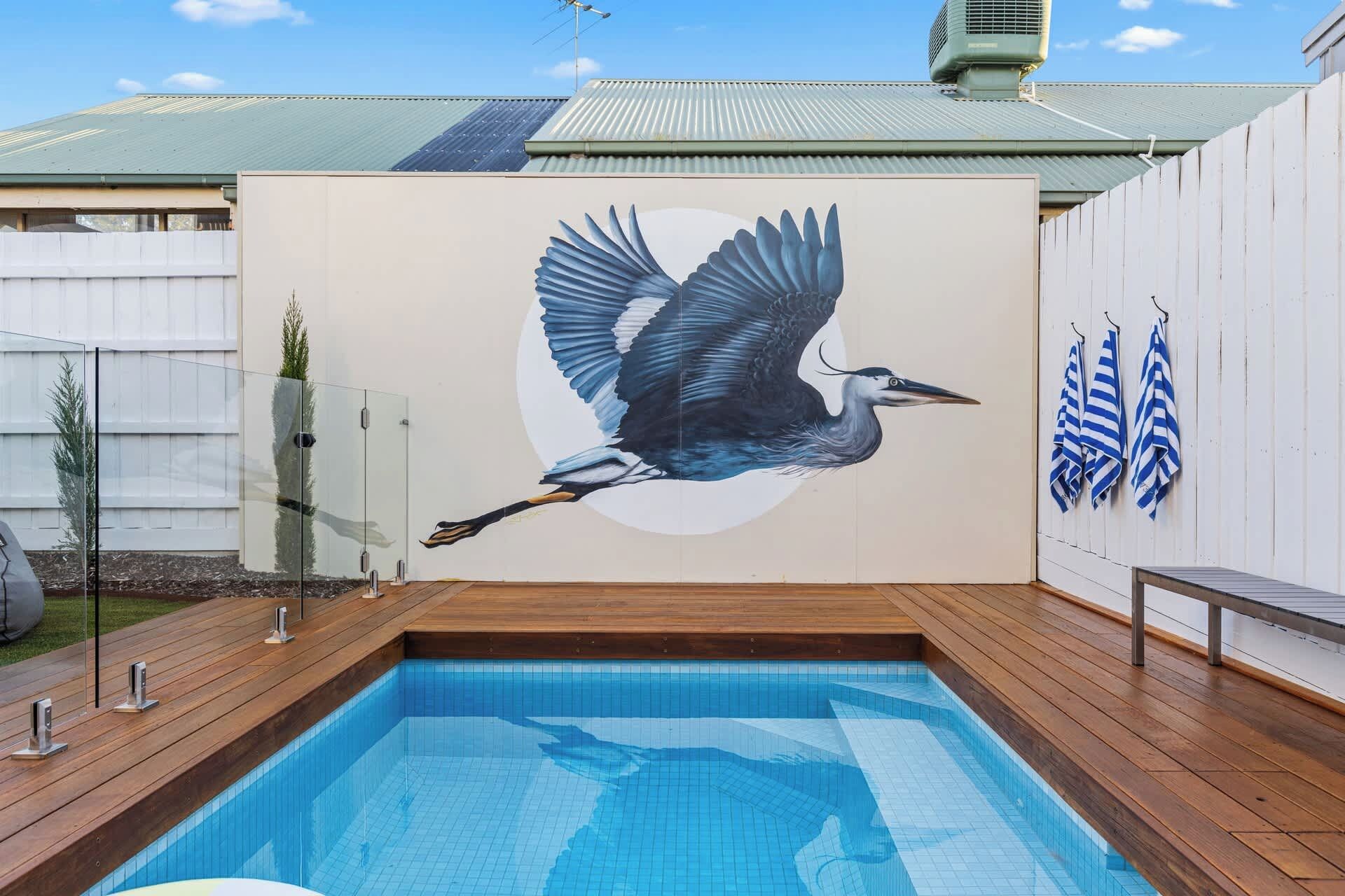 A swimming pool with a mural of a bird in flight, wooden deck, and a white fence.