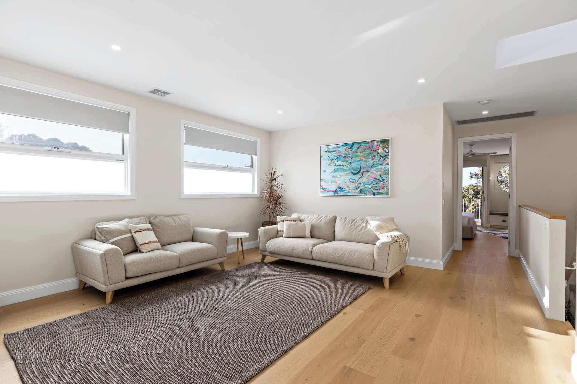 A spacious living room with a large window, a beige sofa, a small round table, and a colorful abstract painting on the wall.