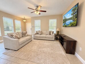 A living room with two sofas, a ceiling fan, a TV, and a painting of a lake. - Family-Friendly Luxury Villa | Sleeps 14 | Private Pool, Game Room & More! (Davenport)