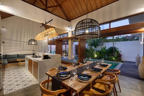 A modern dining area with a long wooden table, multiple chairs, and hanging pendant lights. - Villa Pawikan (Keroboken Kelod)