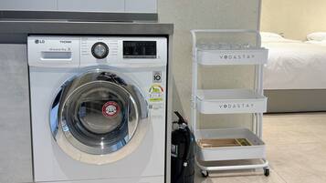 Una secadora LG TROMM de 9 kg con carga frontal y panel de control, un carro de almacenamiento blanco con la marca 'VODASTAY', y un aspirador de pórtatil negro.