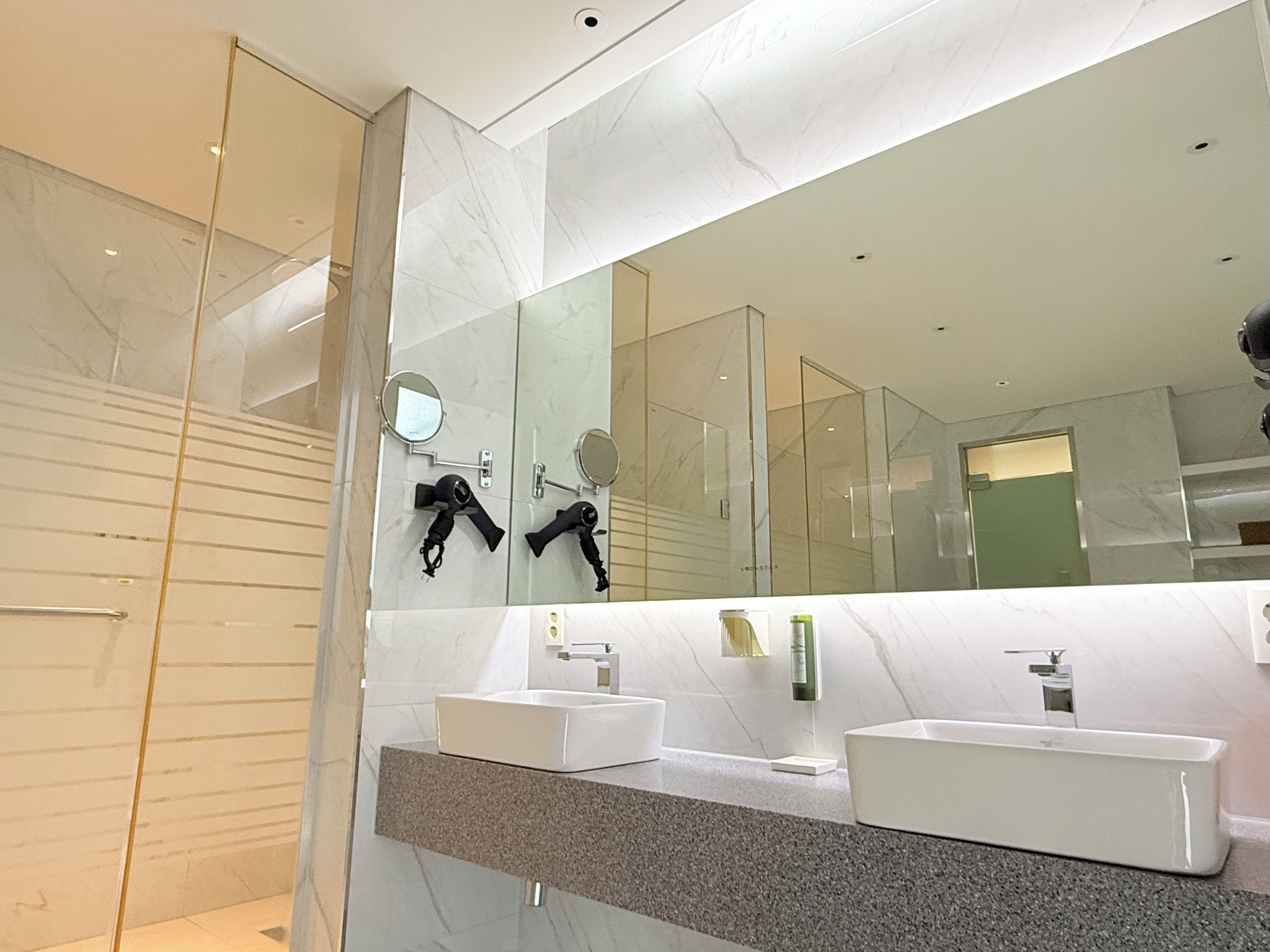 Un baño moderno con un espejo grande, dos lavamanos y una cabina de ducha de vidrio.