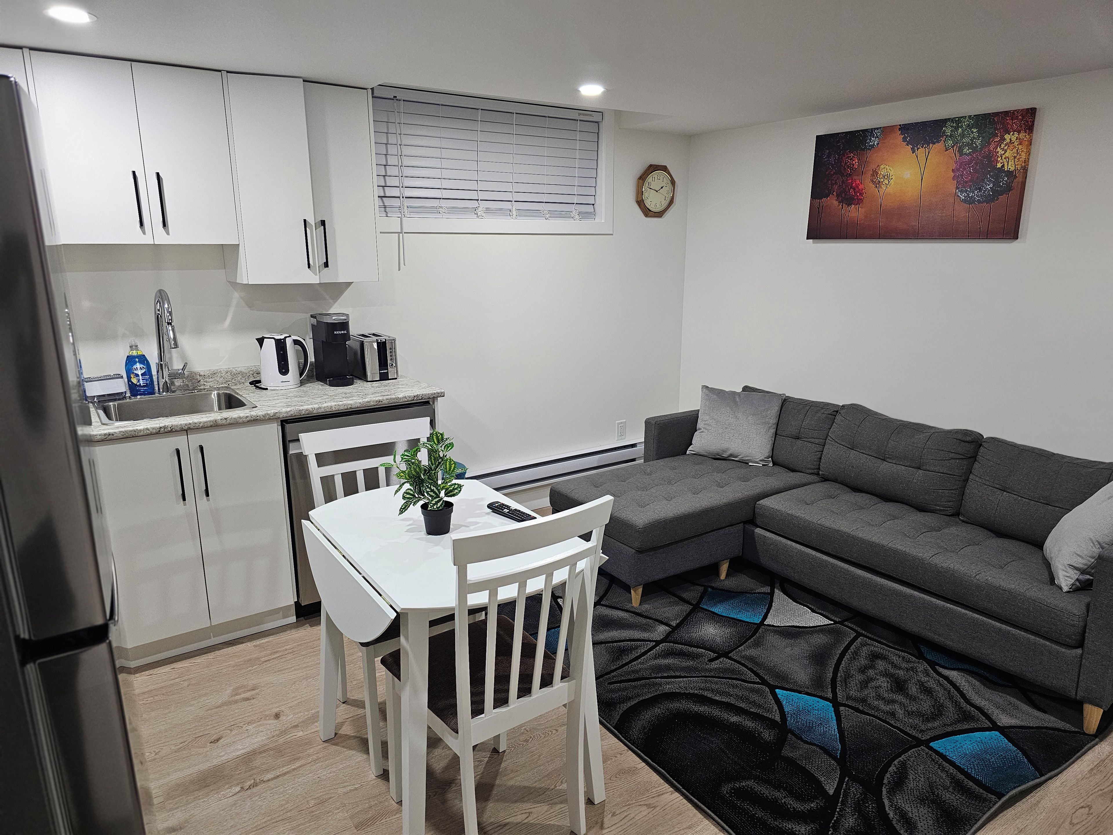 A modern kitchen with white cabinets, a dining table with chairs, a gray sofa, and a colorful rug.