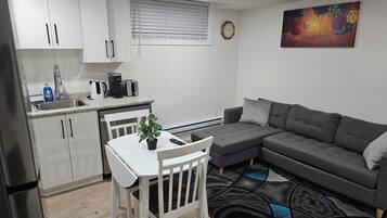 A modern kitchen with white cabinets, a dining table with chairs, a gray sofa, and a colorful rug.