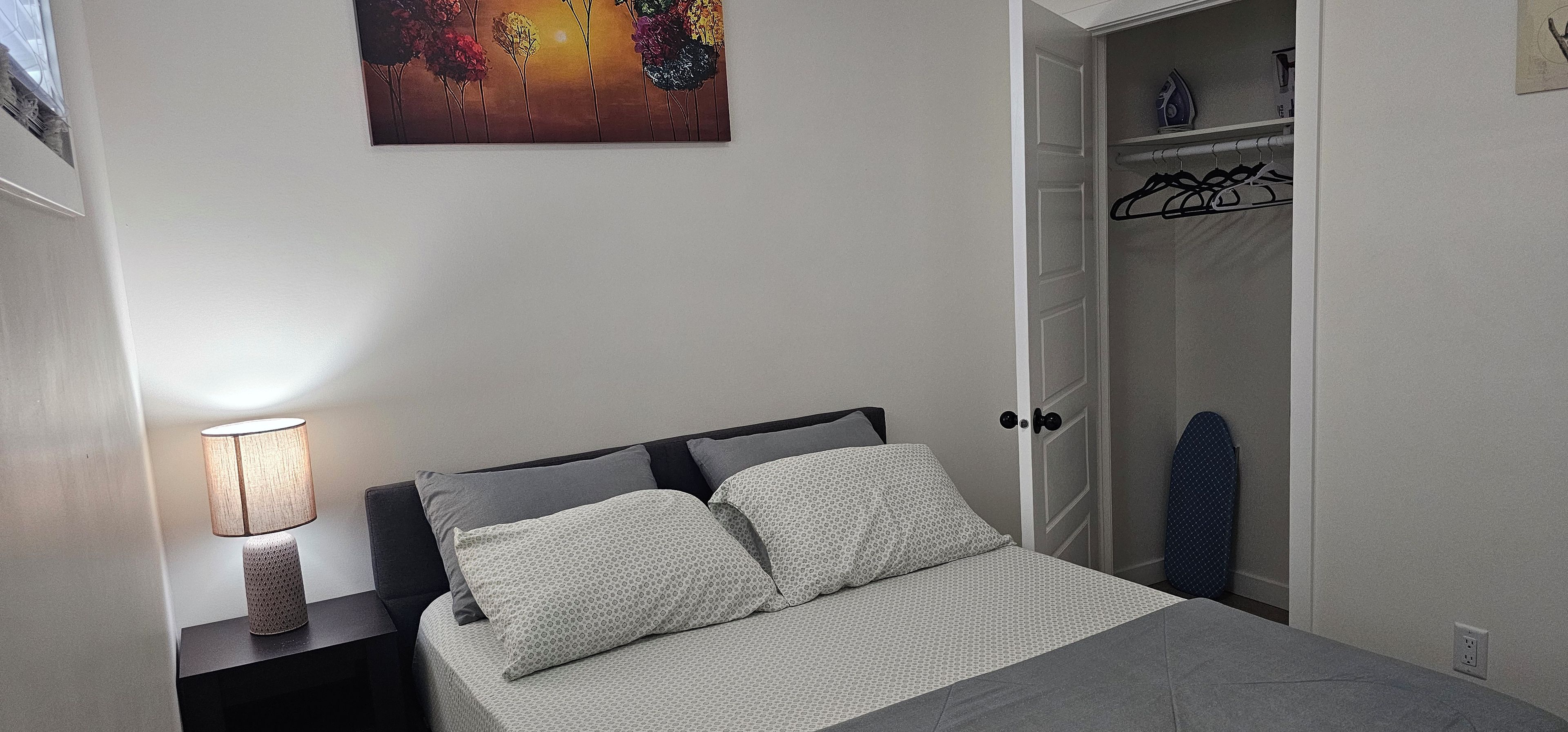 A bedroom with a bed, a nightstand, a lamp, a door, and a closet with hanging clothes.