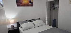 Ein Schlafzimmer mit einem Bett, einem Nachttisch, einer Lampe, einer Tür und einem Kleiderschrank mit Kleidung.