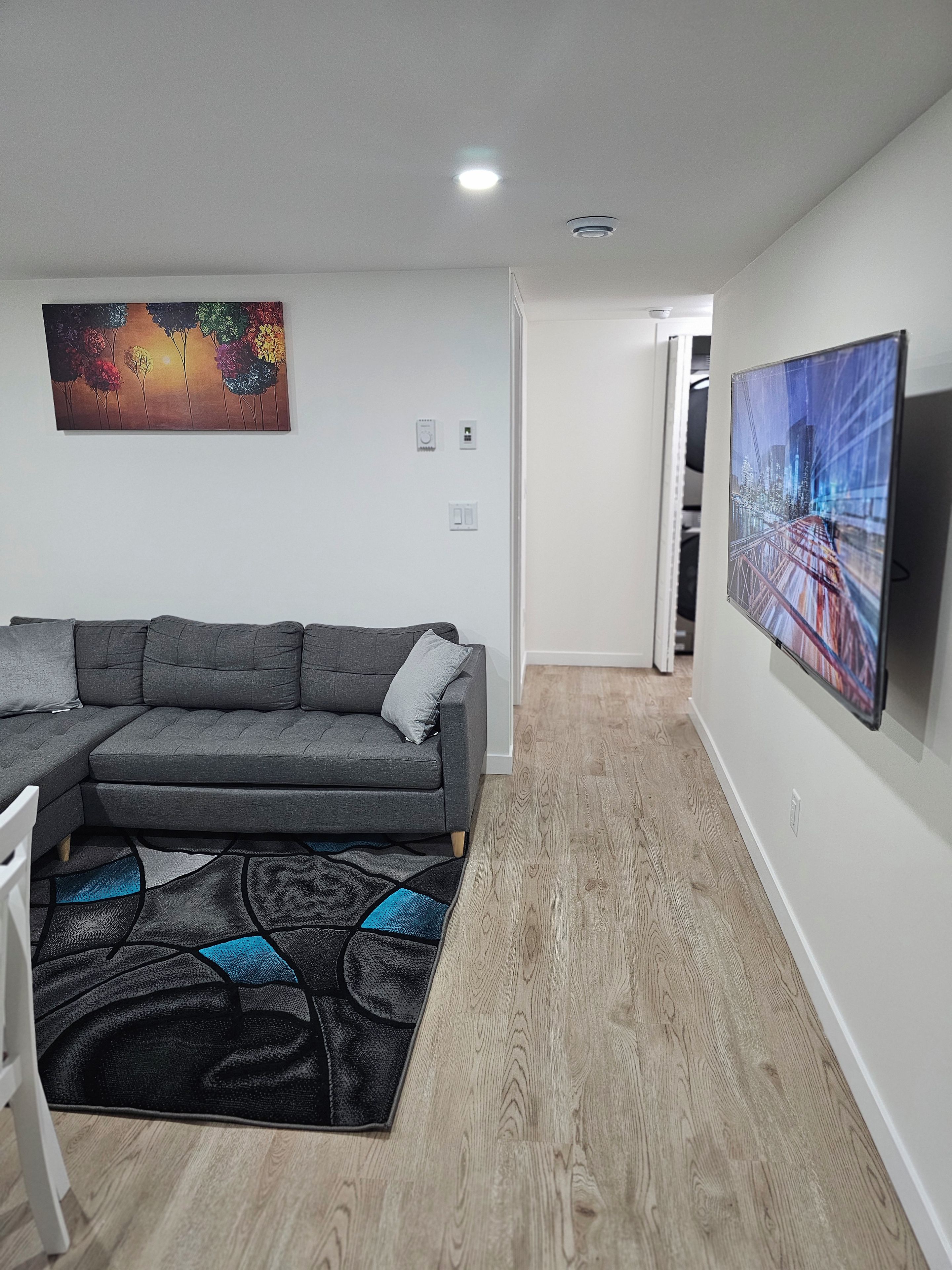 Een moderne woonkamer met een hoekbank, een flatscreen-tv aan de muur en een kleurrijk abstract schilderij erboven.