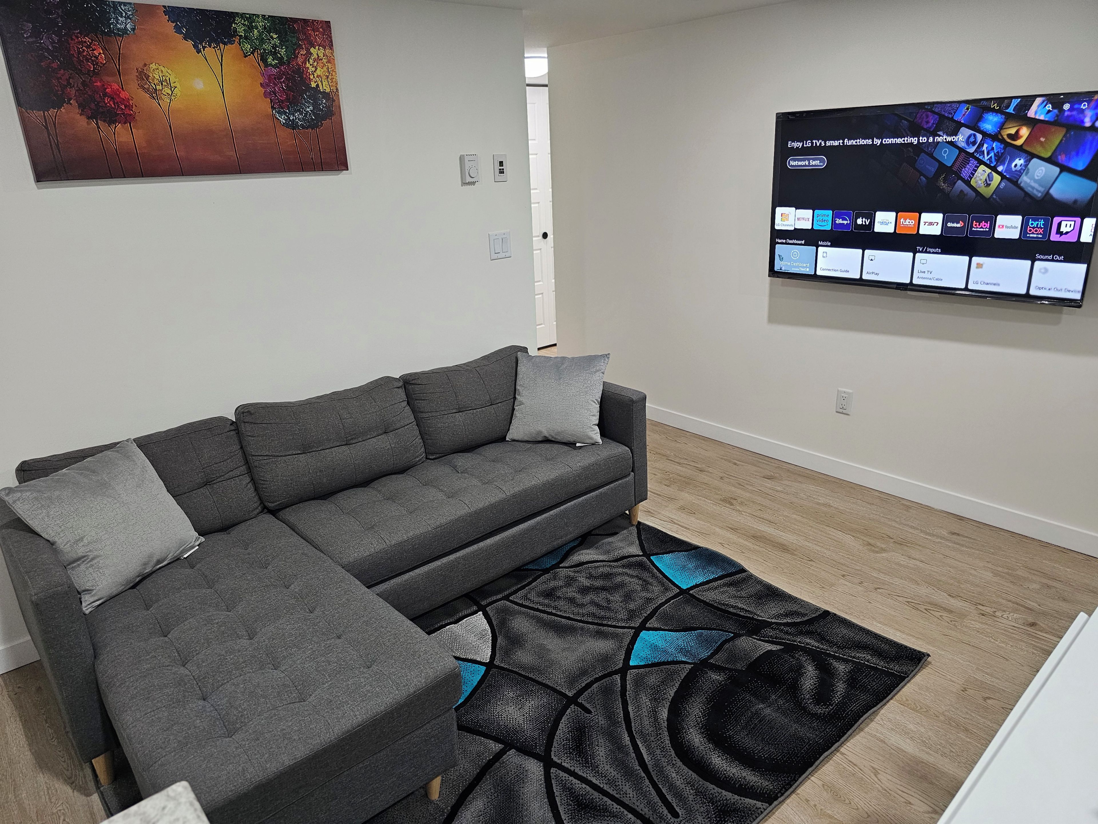 A modern living room with a grey sectional sofa, a patterned rug, a wall-mounted TV displaying various apps, and a colorful painting above the sofa.