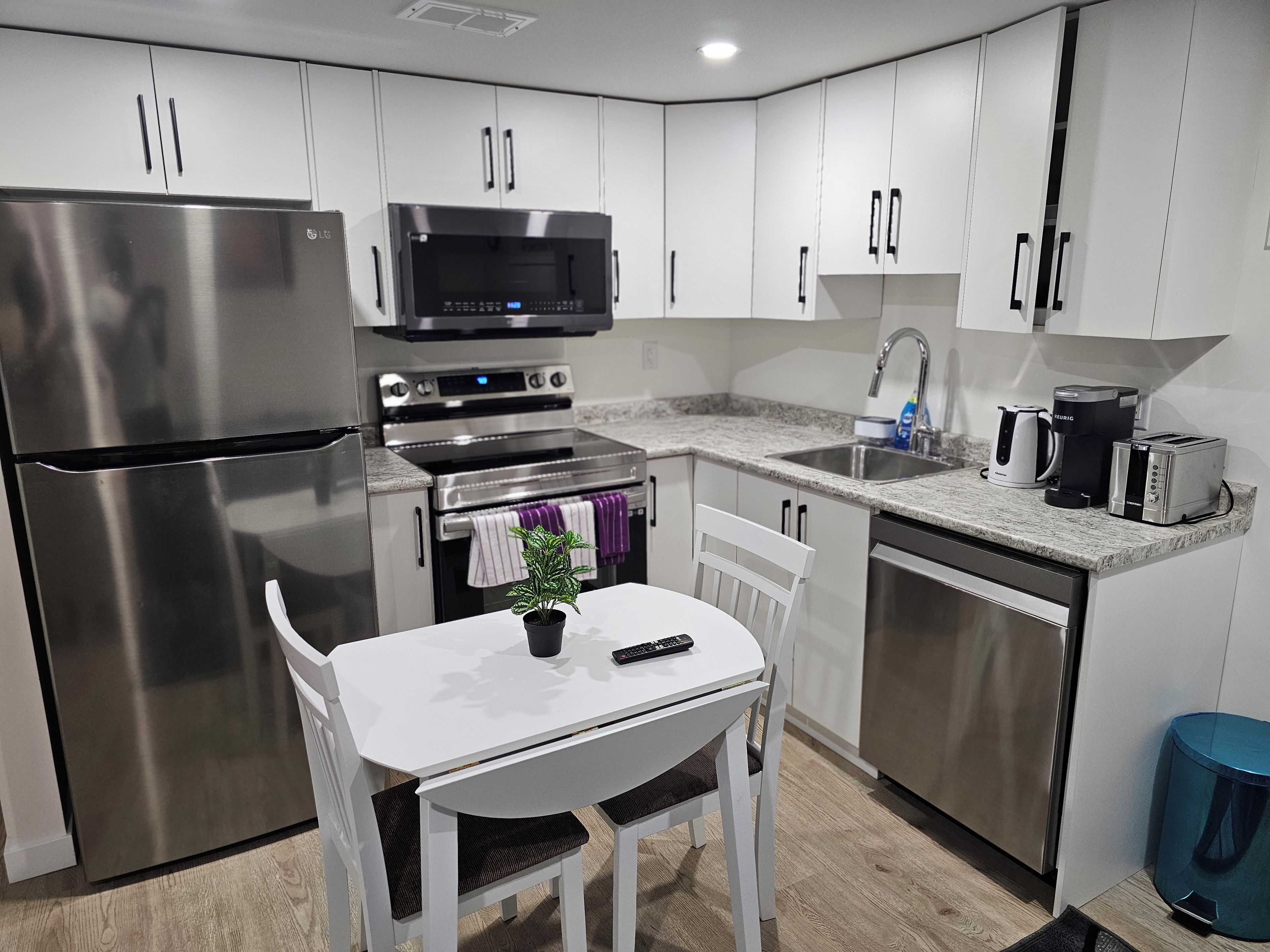 A modern kitchen with stainless steel appliances, a dining table with a chair, and a blue trash bin.