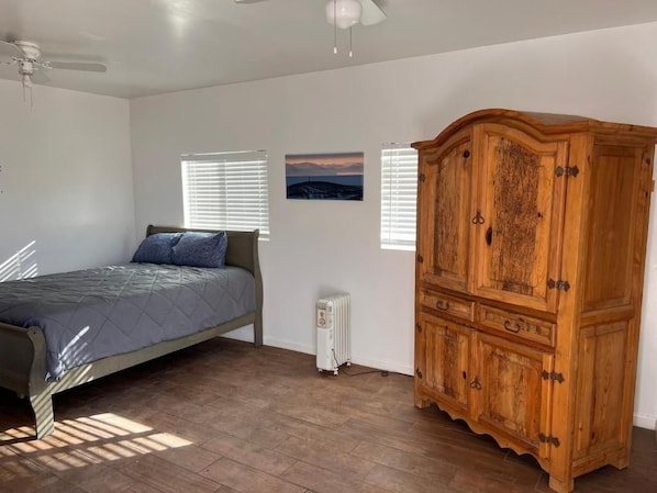 A bedroom with a bed, a radiator, a wooden cabinet, and two windows with blinds. - Moreland Guesthouse (Phoenix)