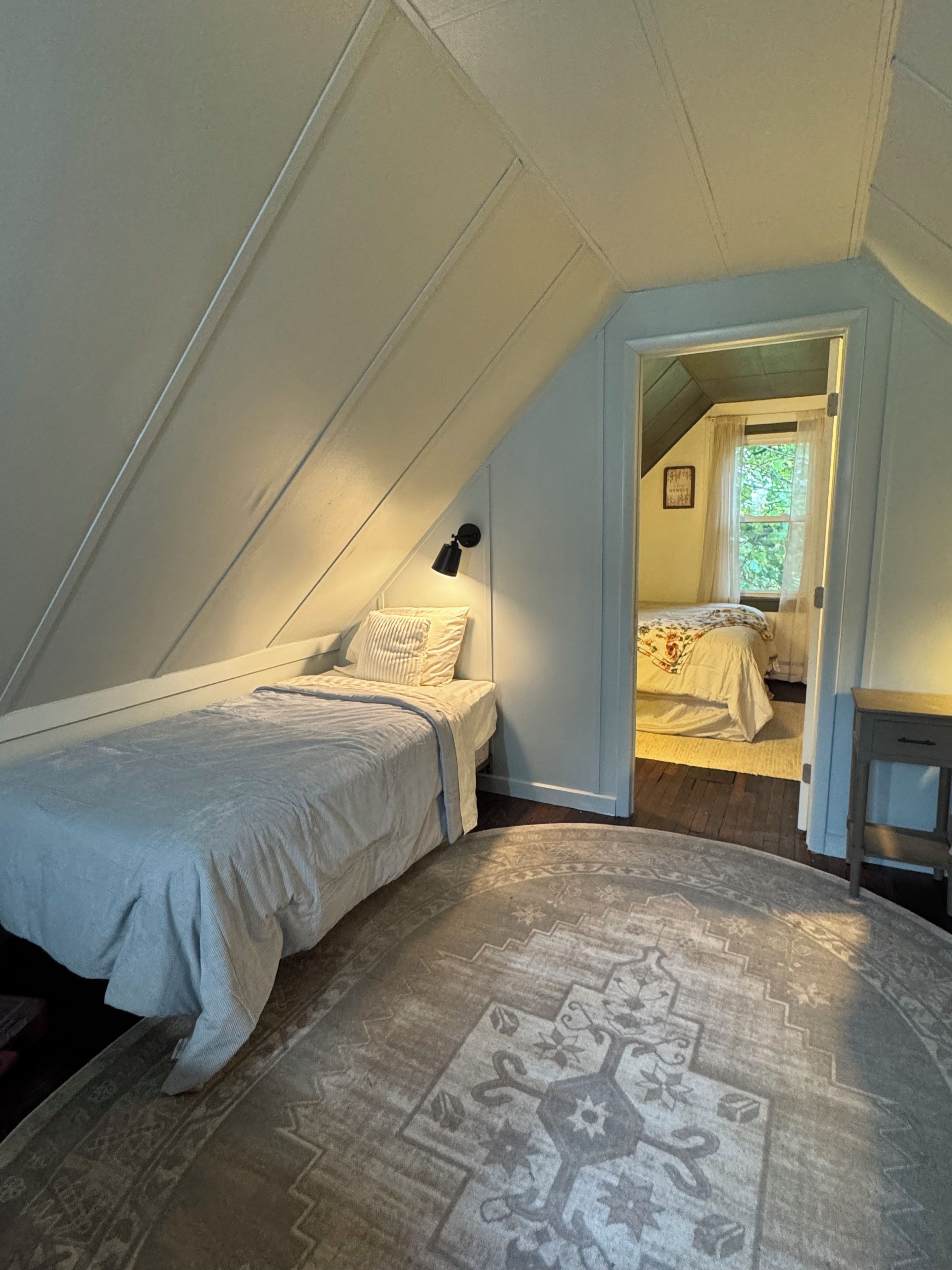 Une chambre avec un plafond en pente, un lit recouvert d’une couette blanche, une lampe de chevet et un tapis à motifs.