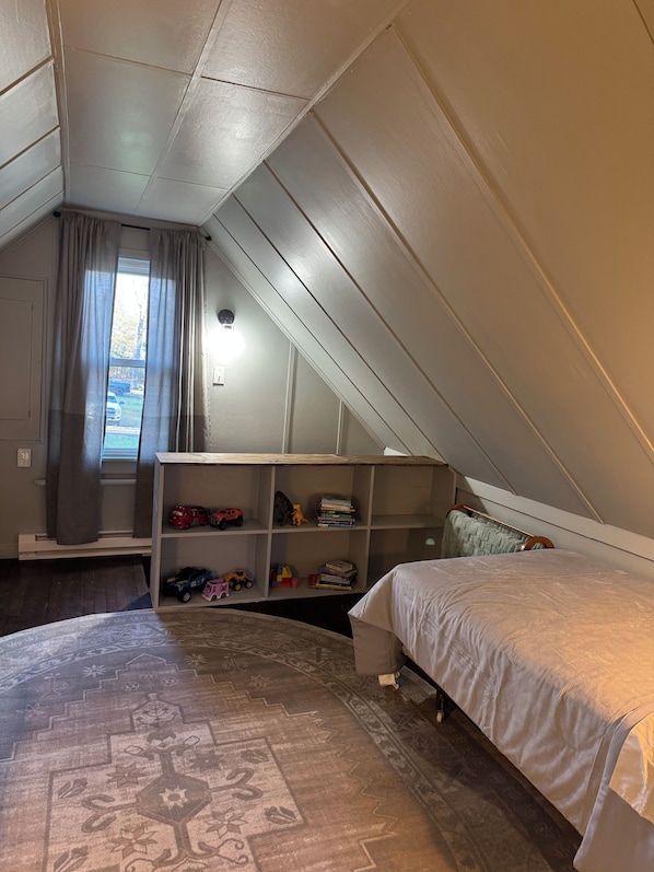 A bedroom with a sloped ceiling, a bed, a bookshelf, and a window with curtains.